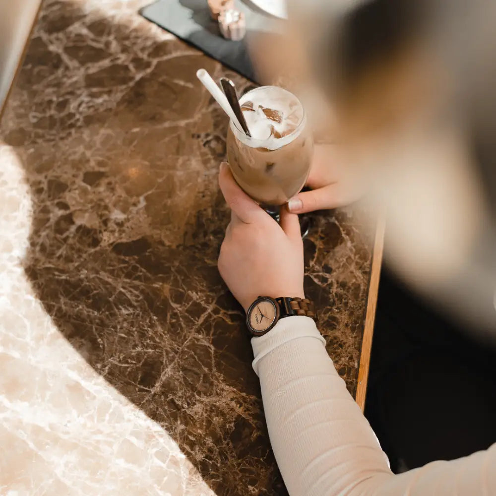 Aava 42mm -rannekello käsissä, jonka edessä on lasi jääkahvia kermavaahdolla ja suklaarouheella
