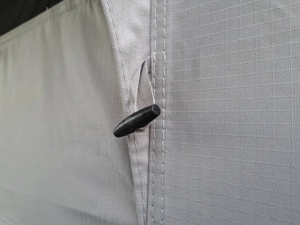 Black plastic toggle clip attached to light gray grid-patterned fabric on auton kattoteltta ilma pitkä 1.6 harmaa