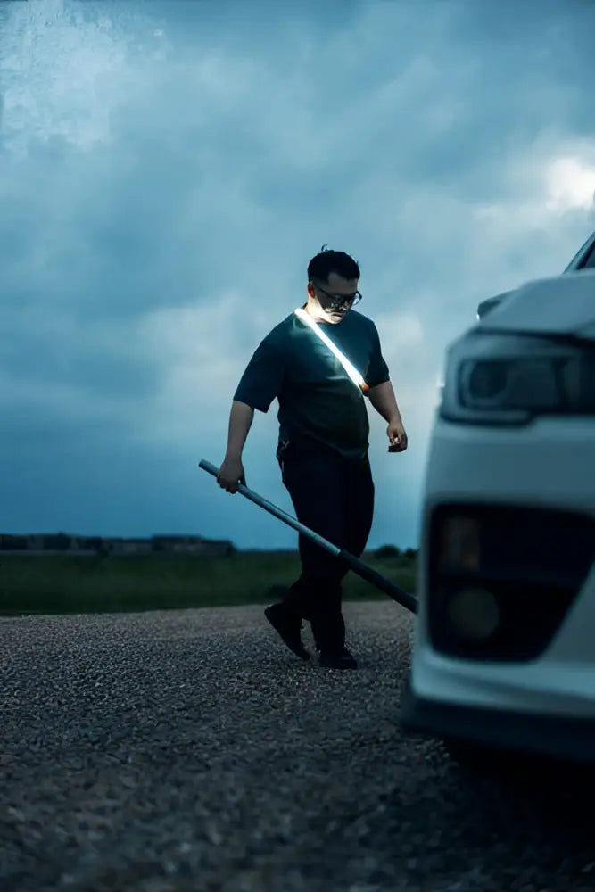 Mies kävelee valkoisen auton vieressä pitäen olallaan valaistua bando 1k strap light työvalon pitkää tankoa