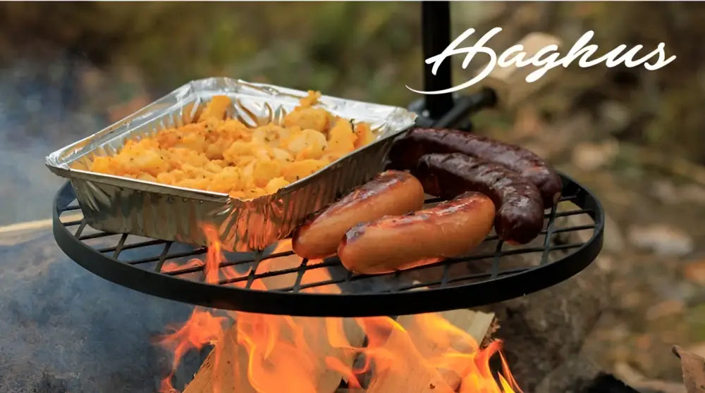 Musta metallinen haghus nuotioteline bbq grillillä ja folioruukku makaronilaatikkoa tulen päällä
