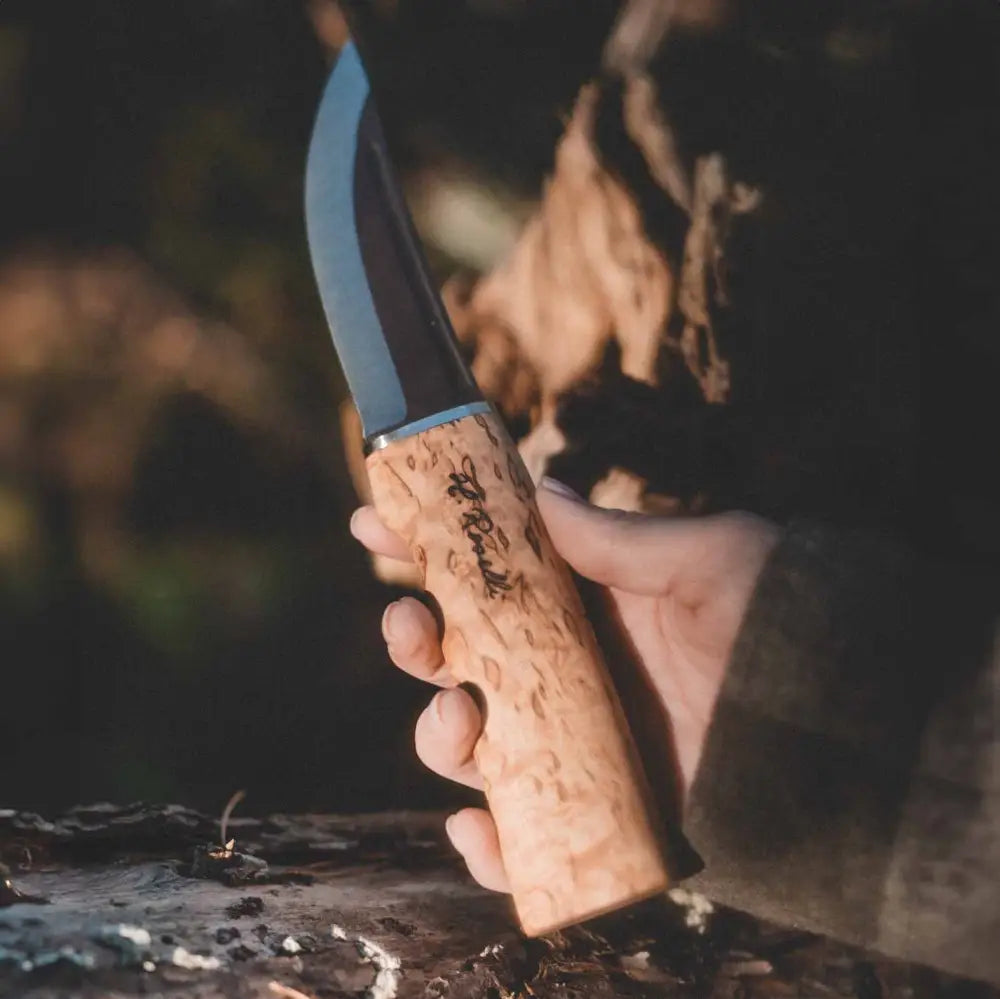 A hand grips a metsästyspuukko knife with a curved polished steel blade and a light brown cork handle with engravings