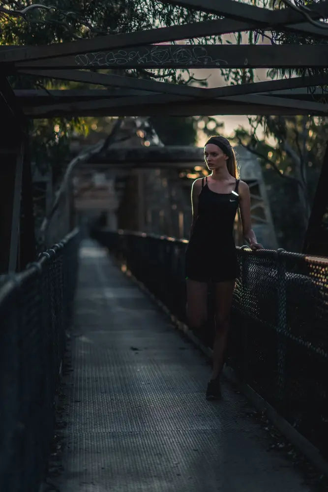 A woman walks along a dimly lit bridge wearing a sleek black sleeveless plus free front dress